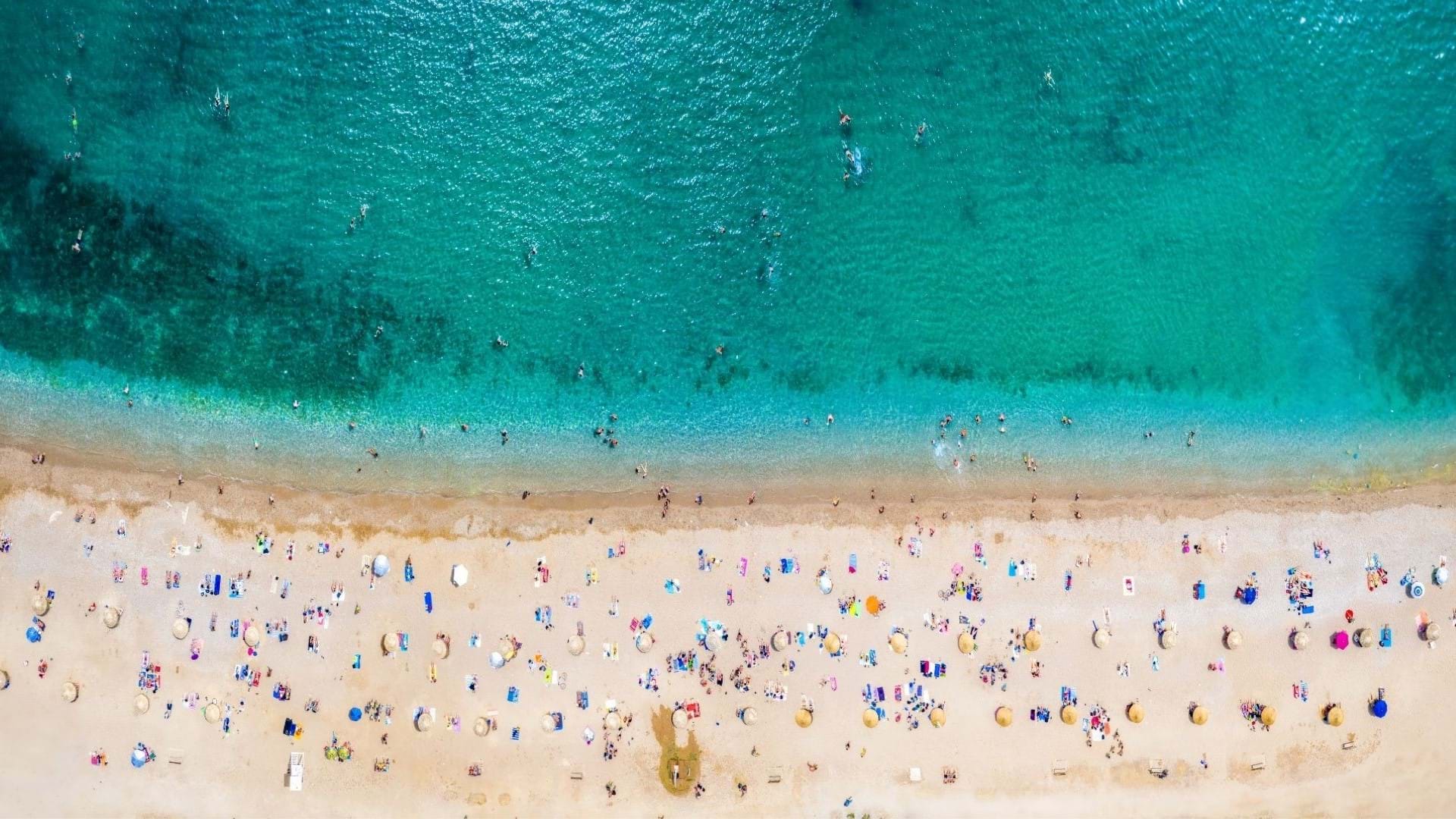 Plaža u Glifadi u Grčkoj, slikana iz ptičje perspektive sa svetlo plavom bojom mora i sitnim peskom i kupačima na plaži.