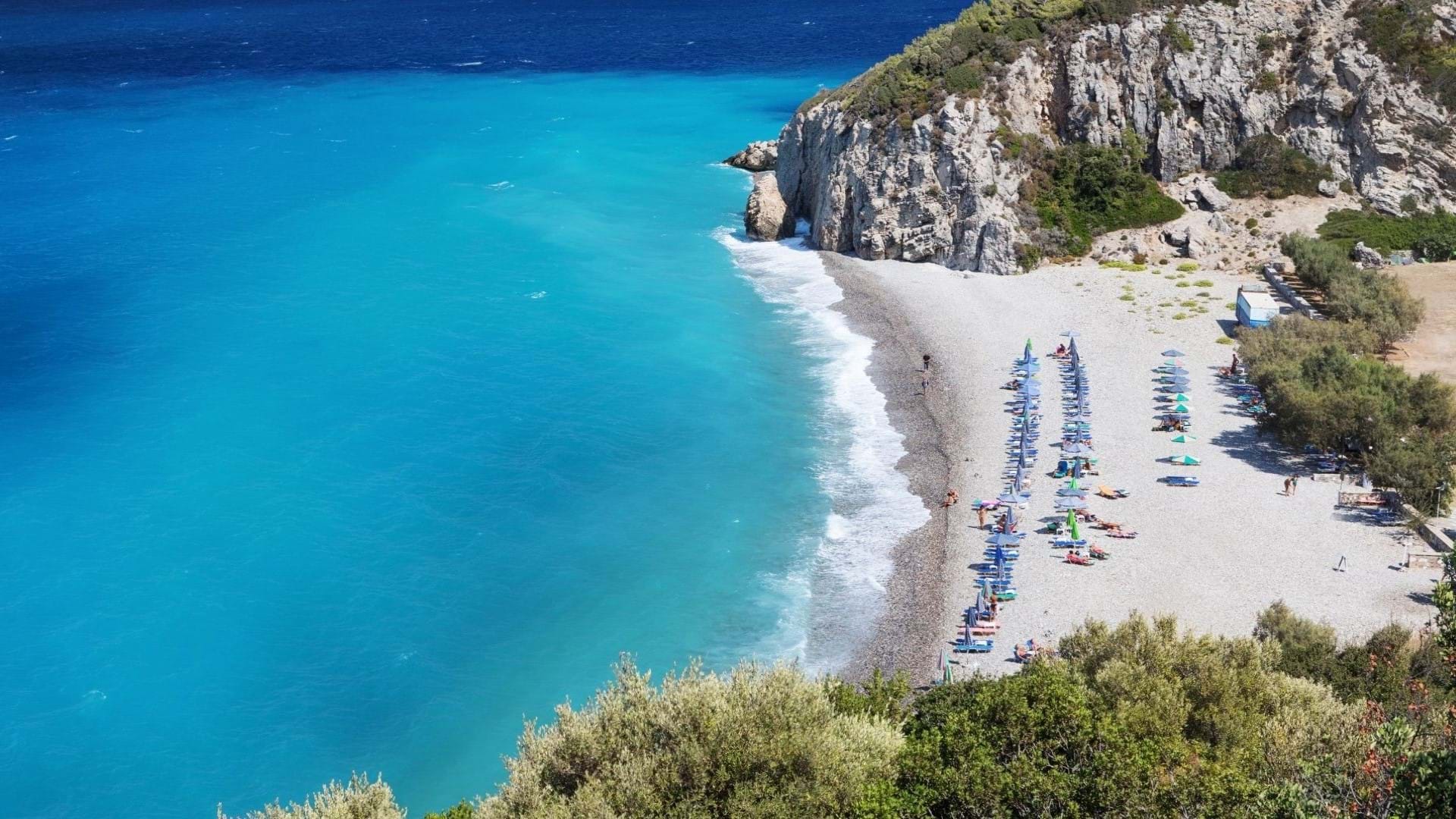 Plaža na Samosu. Šljunkovita obala okružena maslinama, prelepa boja mora i talasi koji zapljuskuju sitan šljunak.